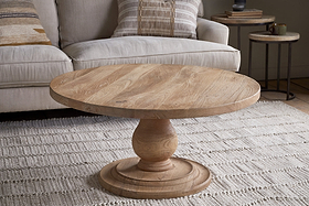 A nkuku Laya Round Carved Coffee Table - Natural sits on a textured cream rug in front of a light beige sofa with patterned cushions and a nested side table in this cozy living room setting.