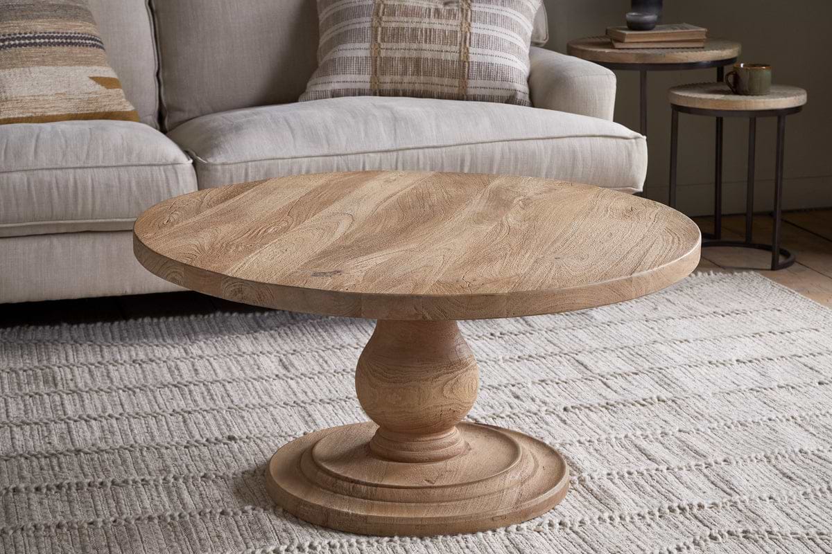 A nkuku Laya Round Carved Coffee Table - Natural sits on a textured cream rug in front of a light beige sofa with patterned cushions and a nested side table in this cozy living room setting.