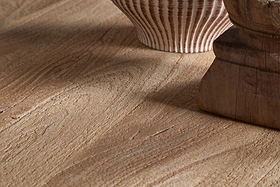 Close-up of the nkuku Laya Round Carved Coffee Table - Natural, showcasing its raw wood finish with distinctive grain and warm brown tones. A ceramic vase and sculptural wooden object appear partially in view.