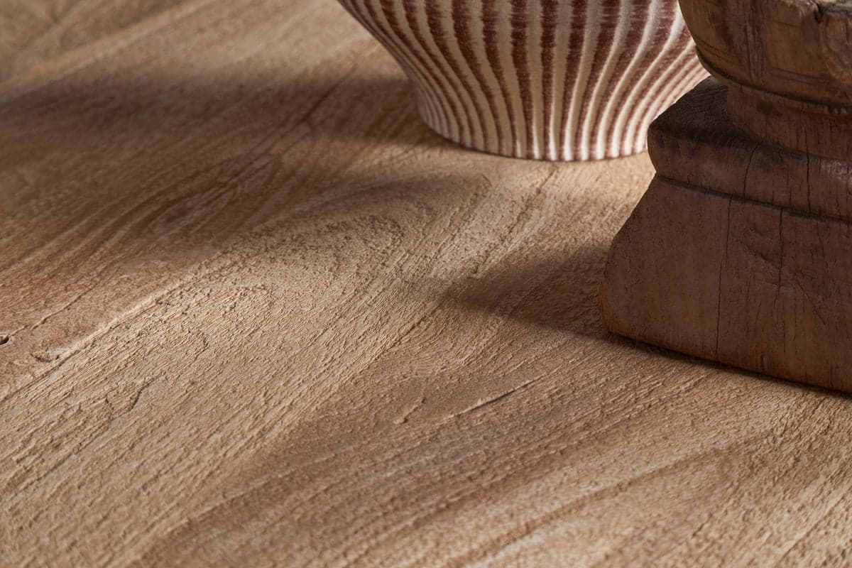 Close-up of the nkuku Laya Round Carved Coffee Table - Natural, showcasing its raw wood finish with distinctive grain and warm brown tones. A ceramic vase and sculptural wooden object appear partially in view.