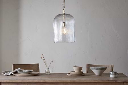 A minimalist dining table with wooden chairs is set with ceramic dinnerware and a linen napkin. On the table stands a small vase of dried flowers. Above, nkuku’s Lumina Fluted Recycled Glass Pendant in clear hangs against a textured white wall.