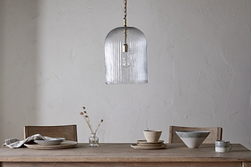 A minimalist dining table with two wooden chairs is set with plates, bowls, a napkin, and a vase of dried flowers. Overhead hangs the nkuku Lumina Fluted Recycled Glass Pendant in clear against a plain textured wall.
