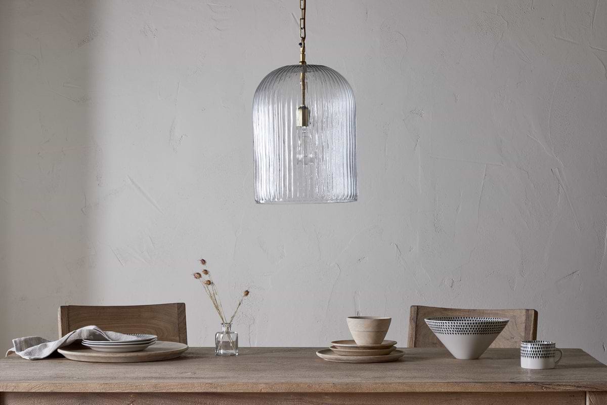 A minimalist dining table with two wooden chairs is set with plates, bowls, a napkin, and a vase of dried flowers. Overhead hangs the nkuku Lumina Fluted Recycled Glass Pendant in clear against a plain textured wall.