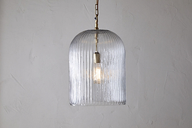 The nkuku Lumina Fluted Recycled Glass Pendant - Clear features a ribbed, dome-shaped shade and brass fixture with a rust-colored cable, casting a gentle glow against a light textured wall.