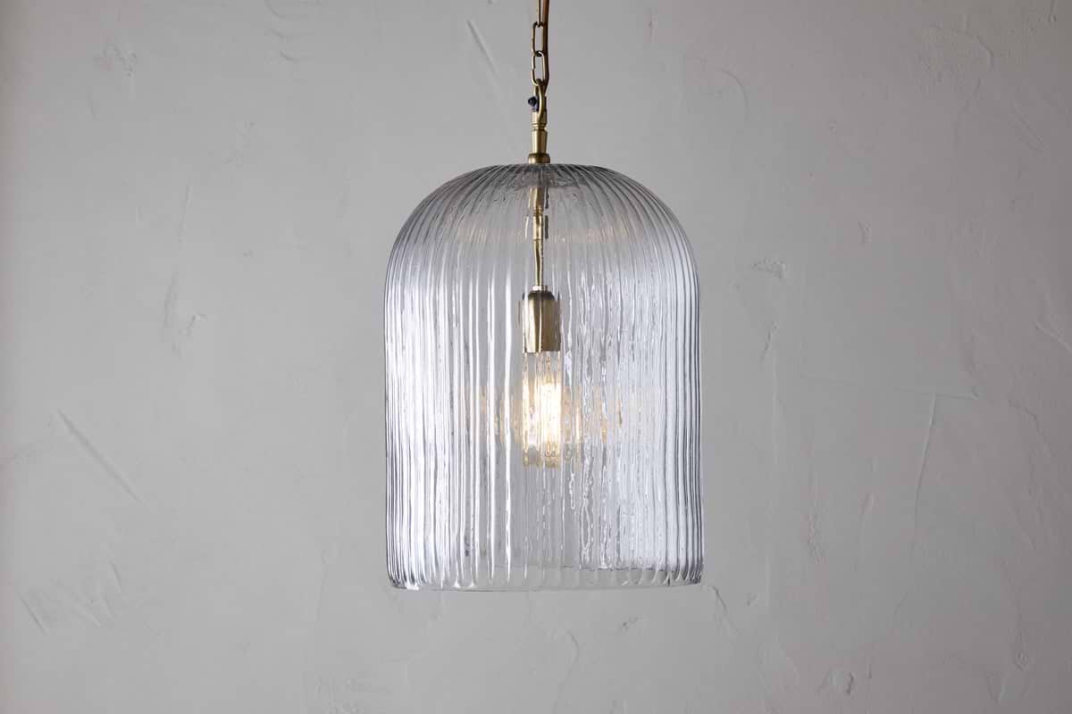 The nkuku Lumina Fluted Recycled Glass Pendant - Clear features a ribbed, dome-shaped shade and brass fixture with a rust-colored cable, casting a gentle glow against a light textured wall.