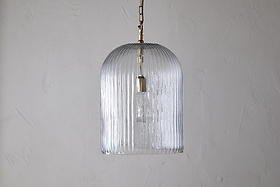 The nkuku Lumina Fluted Recycled Glass Pendant in clear features a ribbed, transparent shade and a rust-coloured fabric cable hanging from a brass fixture, showcasing the bulb against a textured white wall.