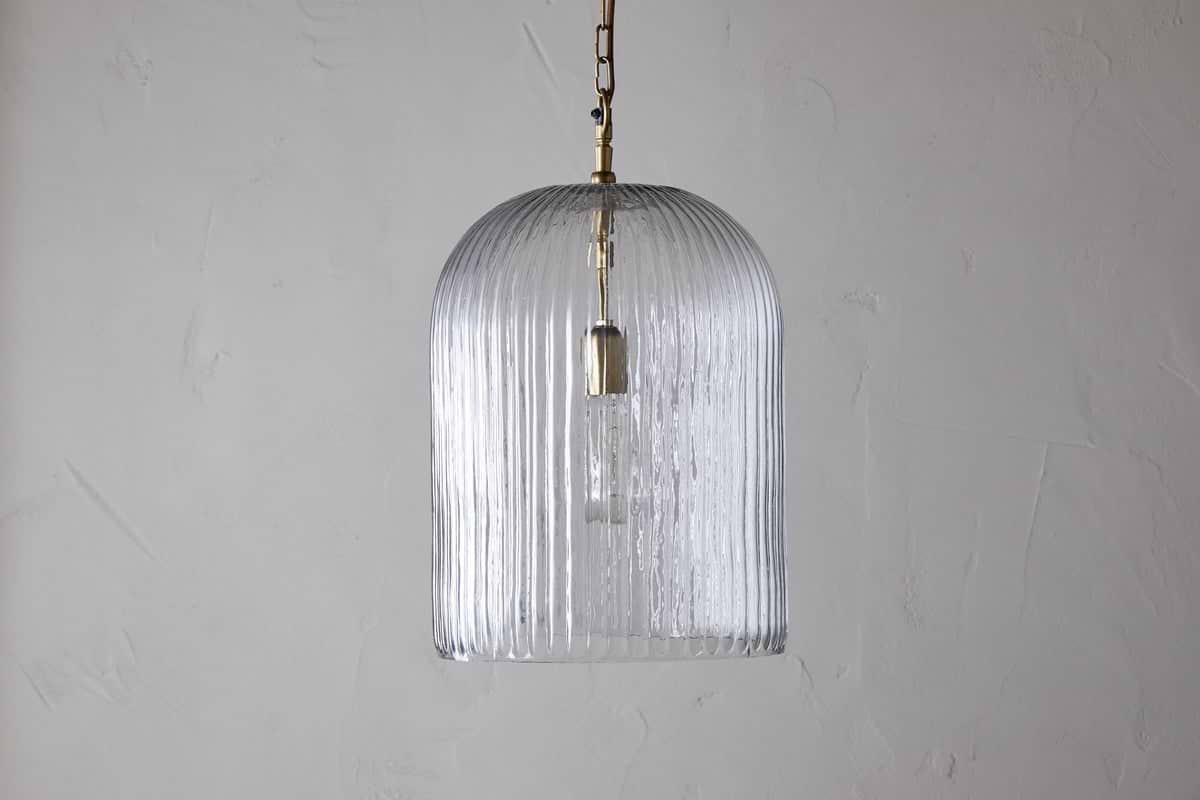 The nkuku Lumina Fluted Recycled Glass Pendant in clear features a ribbed, transparent shade and a rust-coloured fabric cable hanging from a brass fixture, showcasing the bulb against a textured white wall.