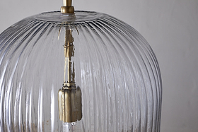 Close-up of the nkuku Lumina Fluted Recycled Glass Pendant in clear, featuring a brass fixture and rust-coloured fabric cable. Vertical grooves accent the transparent shade, with a neutral backdrop highlighting its textured design.
