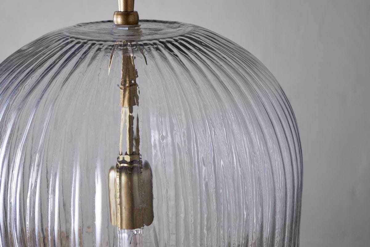 Close-up of the nkuku Lumina Fluted Recycled Glass Pendant in clear, featuring a brass fixture and rust-coloured fabric cable. Vertical grooves accent the transparent shade, with a neutral backdrop highlighting its textured design.