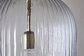 Close-up of the Lumina Fluted Recycled Glass Pendant - Clear by nkuku. The vertical lines and metallic fixture inside stand out against a neutral background, highlighting the pendant's clear recycled glass texture and detail.