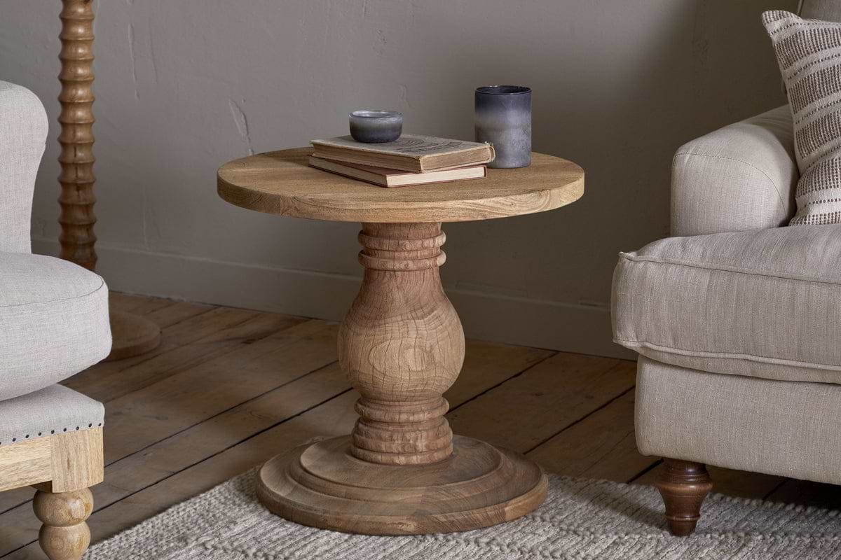 The nkuku Laya Round Carved Side Table - Natural features a sculptural, raw wood design. Shown on a wooden floor between two beige chairs, it holds two books and two small gray cups or candle holders.