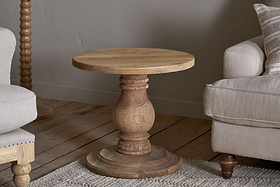 The nkuku Laya Round Carved Side Table in a natural finish sits on a wooden floor between two beige upholstered chairs in a cozy, neutral-toned living room.