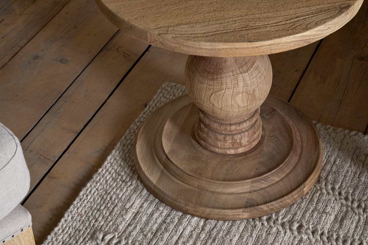 Close-up of the Laya Round Carved Side Table by nkuku, featuring a sculptural pedestal base, shown on a textured beige rug next to a wooden floor and the edge of a light-colored upholstered chair.