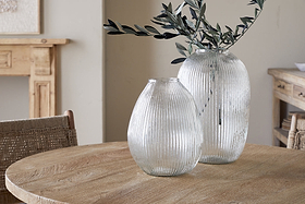Two Linsy Ribbed Glass Vases by nkuku rest on a round wooden table. The larger clear vase displays olive branches, while the smaller is empty. Light wood furniture and a neutral wall add to the calm, rustic atmosphere.