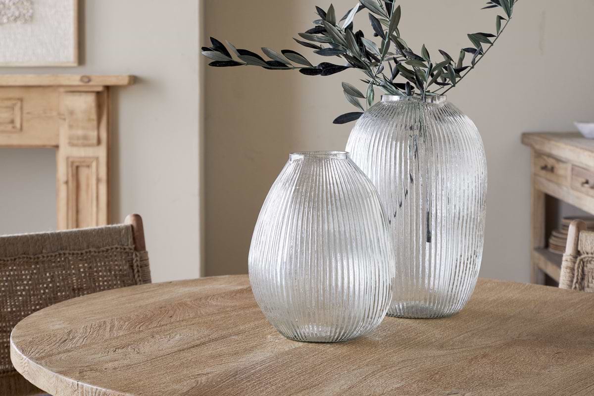 Two Linsy Ribbed Glass Vases by nkuku rest on a round wooden table. The larger clear vase displays olive branches, while the smaller is empty. Light wood furniture and a neutral wall add to the calm, rustic atmosphere.