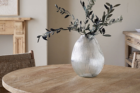 The nkuku Linsy Ribbed Glass Vase - Clear with olive branches stands on a round wooden table. Woven chairs and a light wood sideboard enhance the cozy, minimalist room with neutral walls.