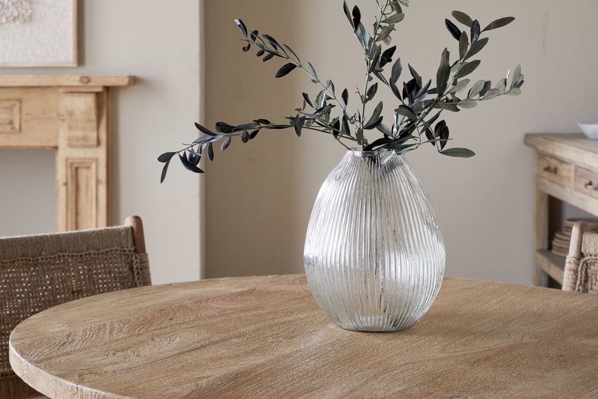 The nkuku Linsy Ribbed Glass Vase - Clear with olive branches stands on a round wooden table. Woven chairs and a light wood sideboard enhance the cozy, minimalist room with neutral walls.
