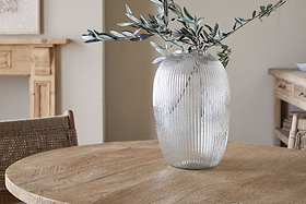 The nkuku Linsy Ribbed Glass Vase - Clear, filled with green olive branches, rests on a round wooden table. Neutral-toned walls, a woven chair, and rustic wood furniture complete the serene, natural interior.