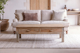 A neutral living room features a light beige sofa, three textured pillows, the nkuku Laali Grooved Mango Wood Coffee Table - Natural with storage drawers, a light woven rug, a console table with decor behind, and a large plant on the left.