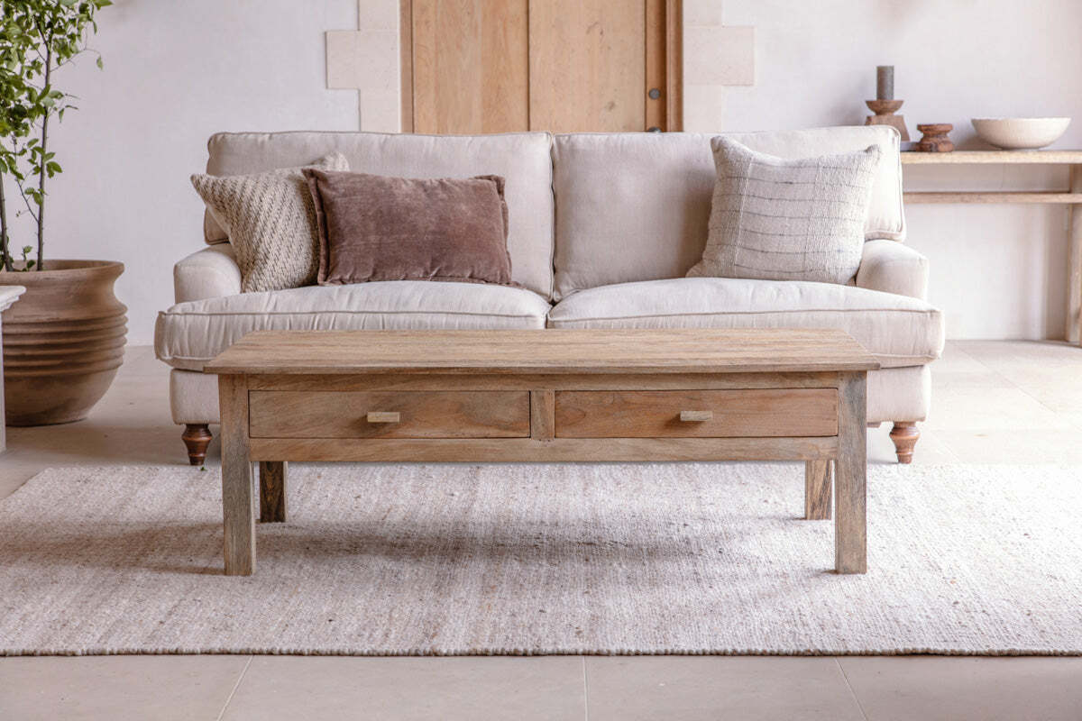 A neutral living room features a light beige sofa, three textured pillows, the nkuku Laali Grooved Mango Wood Coffee Table - Natural with storage drawers, a light woven rug, a console table with decor behind, and a large plant on the left.