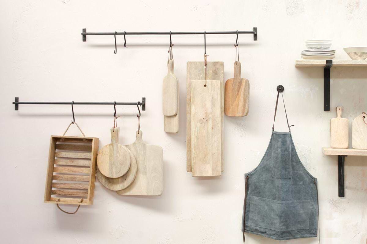 The nkuku Laila Hanging Rail in iron holds wooden cutting boards, a washboard, and an apron on hooks. A nearby wooden shelf with stacked white dishes adds to the organized, stylish look against the light wall in your kitchen.