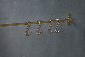 Four Laila Iron S-Hooks in a brass finish by nkuku are evenly spaced on a traditional brass rod, mounted to a textured dark gray wall with a matching right-side bracket; no items hang from the S-hooks.