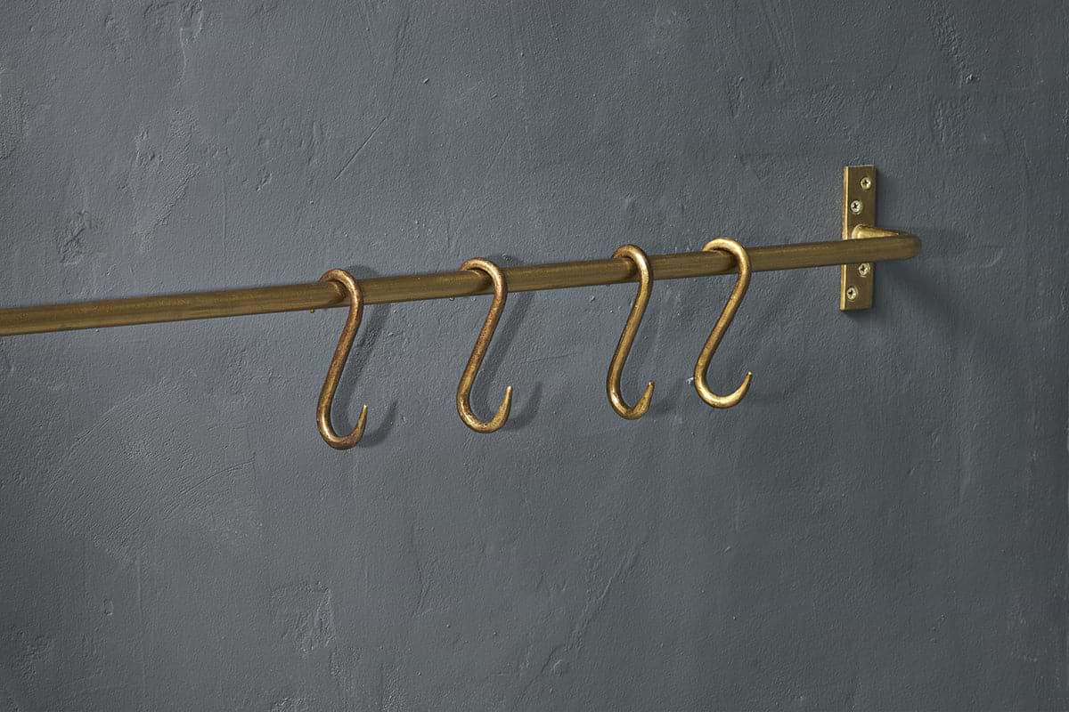 Four Laila Iron S-Hooks in a brass finish by nkuku are evenly spaced on a traditional brass rod, mounted to a textured dark gray wall with a matching right-side bracket; no items hang from the S-hooks.