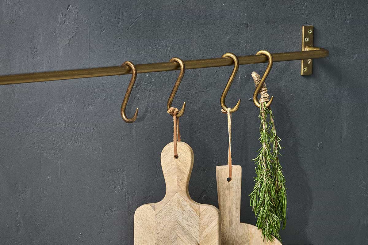 The nkuku Laila Iron S-Hooks in brass (set of 4) hold two light wood cutting boards with leather loops and a bundle of rosemary, all displayed against a matte dark gray textured wall.