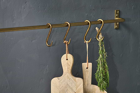 The nkuku Laila Iron S-Hooks in brass (set of 4) hold two light wood cutting boards with leather loops and a bundle of rosemary, all displayed against a matte dark gray textured wall.
