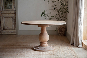 The Laya Round Carved Mango Wood Table - Small by nkuku rests on a woven rug in a softly lit room with wood floors, rustic door, potted plant, and sheer curtains, creating a warm and inviting natural ambiance.
