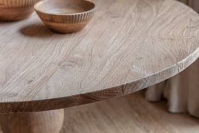 Close-up of the Laya Round Carved Mango Wood Table - Small by nkuku, seating 4. Visible wood grain and two carved wooden bowls emphasize its artisanal craftsmanship.