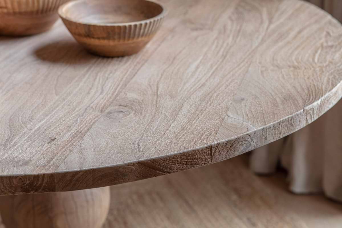 Close-up of the Laya Round Carved Mango Wood Table - Small by nkuku, seating 4. Visible wood grain and two carved wooden bowls emphasize its artisanal craftsmanship.