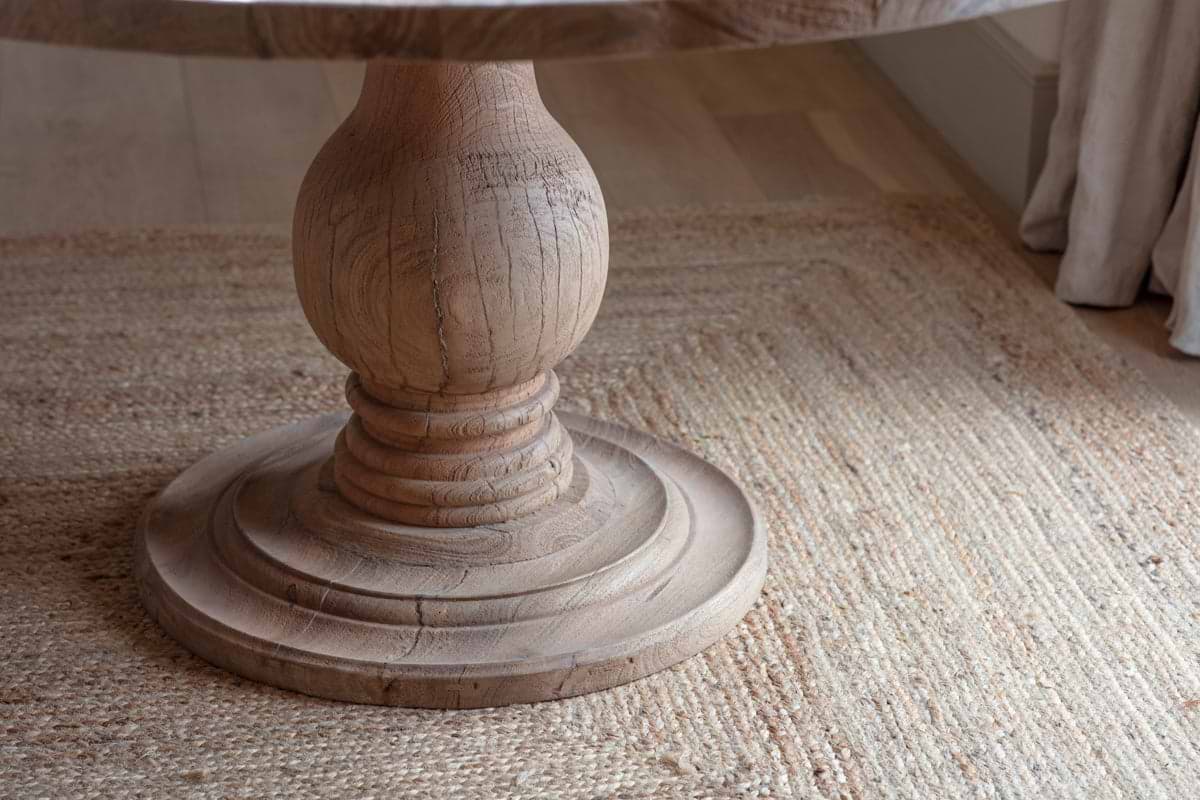A close-up of the Laya Round Carved Mango Wood Table - Small by nkuku, featuring a hand-carved pedestal base with a natural finish, set on a woven light brown rug over light hardwood floors and beige curtains in the background.