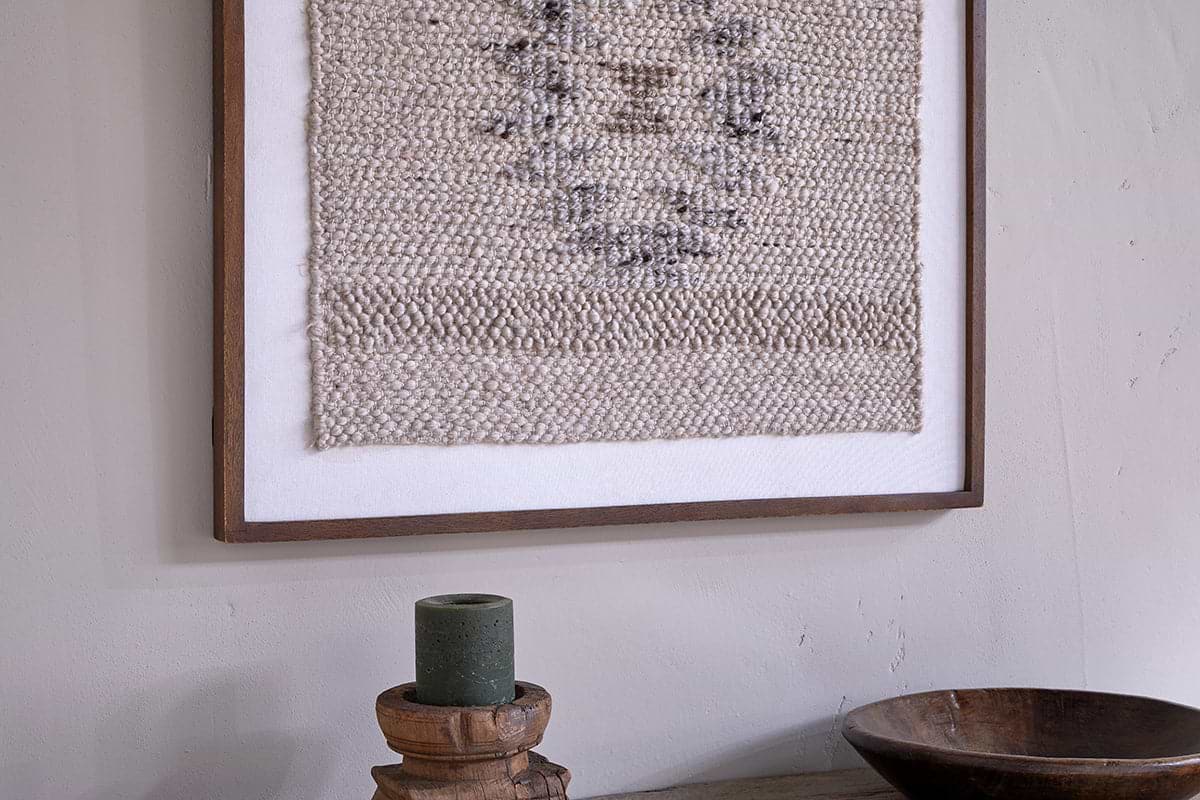 The Lena Textured Wall Art - Off White by nkuku hangs above a rustic wooden surface, where a round wooden bowl and a green candle on a carved stand enhance the space’s natural, earthy vibe.