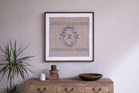 A rustic wooden dresser with three drawers holds a potted plant, candles, and a wooden bowl. Above, the nkuku Lena Textured Wall Art in Off White adds artisanal charm to the light beige wall.