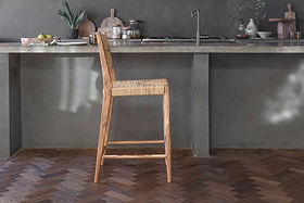 The Lohanda Acacia & Munja Grass Counter Chair by nkuku stands before a gray concrete island in a modern, minimalist kitchen with potted plants, wooden boards, jars, and tableware on dark herringbone wood floors.