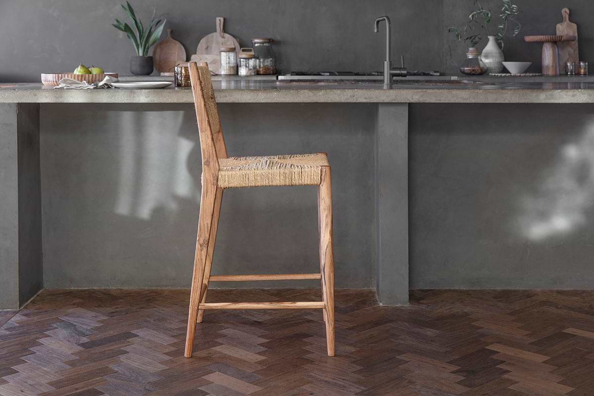 The Lohanda Acacia & Munja Grass Counter Chair by nkuku stands before a gray concrete island in a modern, minimalist kitchen with potted plants, wooden boards, jars, and tableware on dark herringbone wood floors.