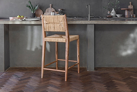 The nkuku Lohanda Acacia & Munja Grass Counter Chair - Natural stands before a gray concrete kitchen counter topped with plates, bowls, chopping boards, and a small plant above dark herringbone wood flooring.