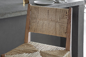Close-up of the nkuku Lohanda Acacia & Munja Grass Counter Chair in natural, featuring a wooden frame and woven seat, next to a grey concrete dining table set with a white plate and cutlery.