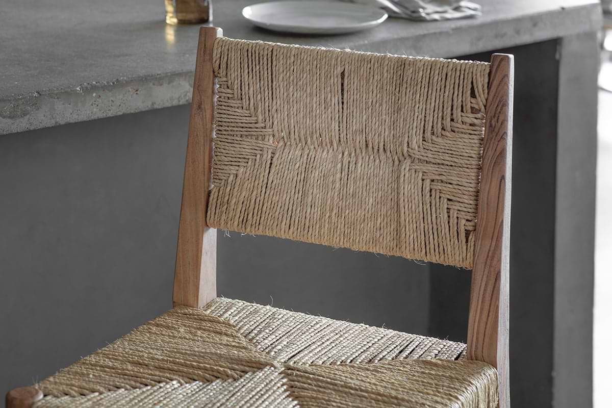 Close-up of the nkuku Lohanda Acacia & Munja Grass Counter Chair in natural, featuring a wooden frame and woven seat, next to a grey concrete dining table set with a white plate and cutlery.