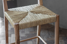 The nkuku Lohanda Acacia & Munja Grass Counter Chair features a handcrafted acacia frame and a seat intricately woven from natural beige munja grass, highlighting artisanal craftsmanship and elegant geometric patterns.