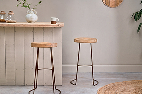 A light wood breakfast bar with two nkuku Loko Mango Wood Stools - Tall, featuring metal legs. On the bar sit a white teapot, mug, glass jars, and a vase with green stems. The room has light walls, a plant, and a round woven rug at the edge.