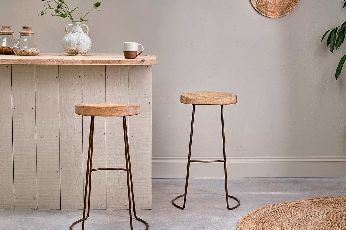 A light wood breakfast bar with two nkuku Loko Mango Wood Stools - Tall, featuring metal legs. On the bar sit a white teapot, mug, glass jars, and a vase with green stems. The room has light walls, a plant, and a round woven rug at the edge.