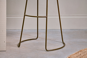 Close-up of the Loko Mango Wood Stool - Tall by nkuku, featuring a modern rustic iron base with thin curved legs and a footrest on a smooth light gray floor, against a white wall with trim and part of a woven rug in the corner.