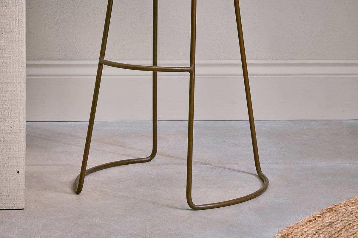 Close-up of the Loko Mango Wood Stool - Tall by nkuku, featuring a modern rustic iron base with thin curved legs and a footrest on a smooth light gray floor, against a white wall with trim and part of a woven rug in the corner.