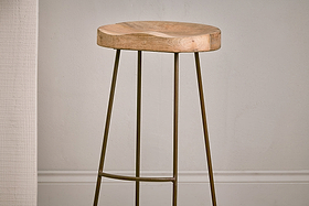 The Loko Mango Wood Stool - Tall by nkuku features a minimalist design with a rounded, smooth mango wood seat and three slender outward-angled metal legs, set against a plain light wall with white trim.