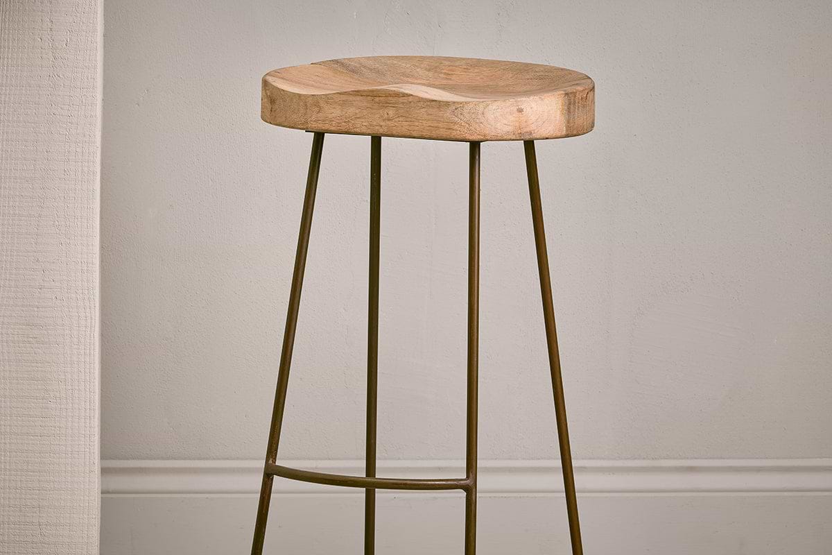 The Loko Mango Wood Stool - Tall by nkuku features a minimalist design with a rounded, smooth mango wood seat and three slender outward-angled metal legs, set against a plain light wall with white trim.