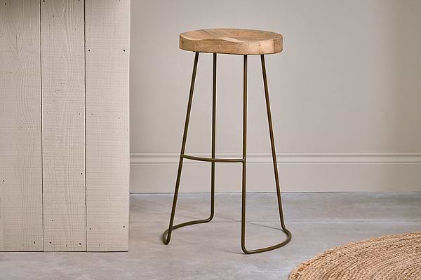 The nkuku Loko Mango Wood Stool - Tall features a round, carved mango wood seat and slim dark metal legs, set on a grey concrete floor beside a light vertical wooden panel.