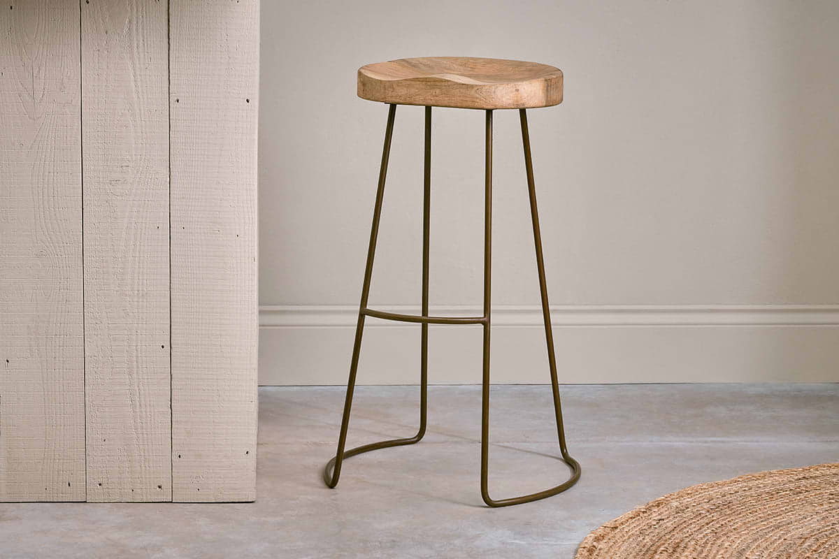 The nkuku Loko Mango Wood Stool - Tall features a round, carved mango wood seat and slim dark metal legs, set on a grey concrete floor beside a light vertical wooden panel.
