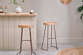 Two Loko Mango Wood Stools by nkuku, with dark iron legs and sustainable mango wood seats, stand in front of a light wood bar. A textured white vase with greenery and a two-tone mug complete the inviting scene.